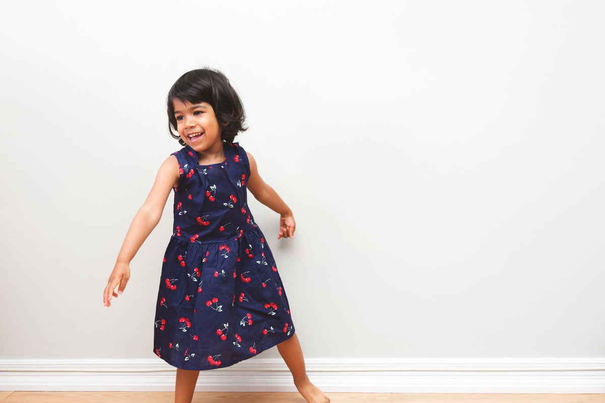 An older toddler girl smiling and playing wearing a dark blue dress in front of a white wall.